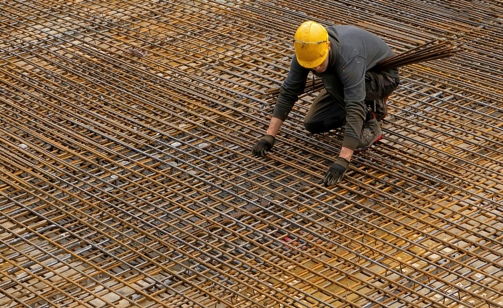 Worker in a yellow hard hat kneeling to tie a rusty rebar mat before a pour