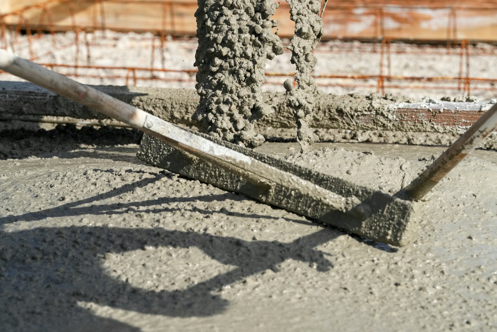 Close-up of a hand screed leveling freshly placed concrete while more concrete pours in behind it