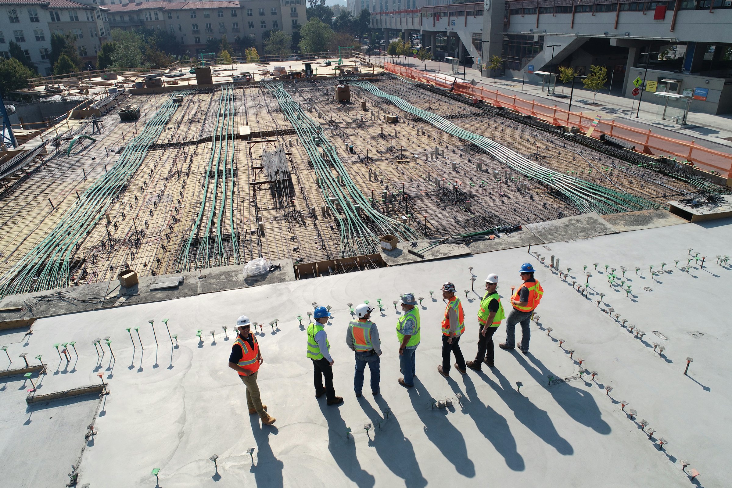 Overhead view of a seven-person crew in hard hats and hi-vis vests standing on a finished concrete deck, next to an unpoured bay of rebar and conduit