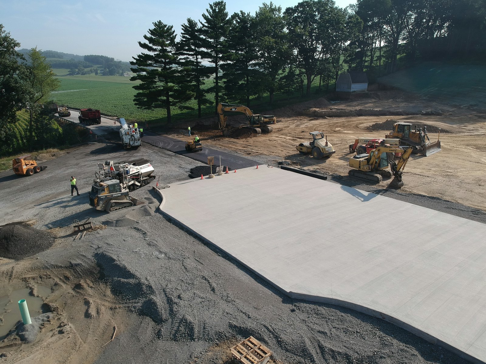 Aerial view of a large, freshly placed concrete slab on a graded site with paving equipment and a tree line