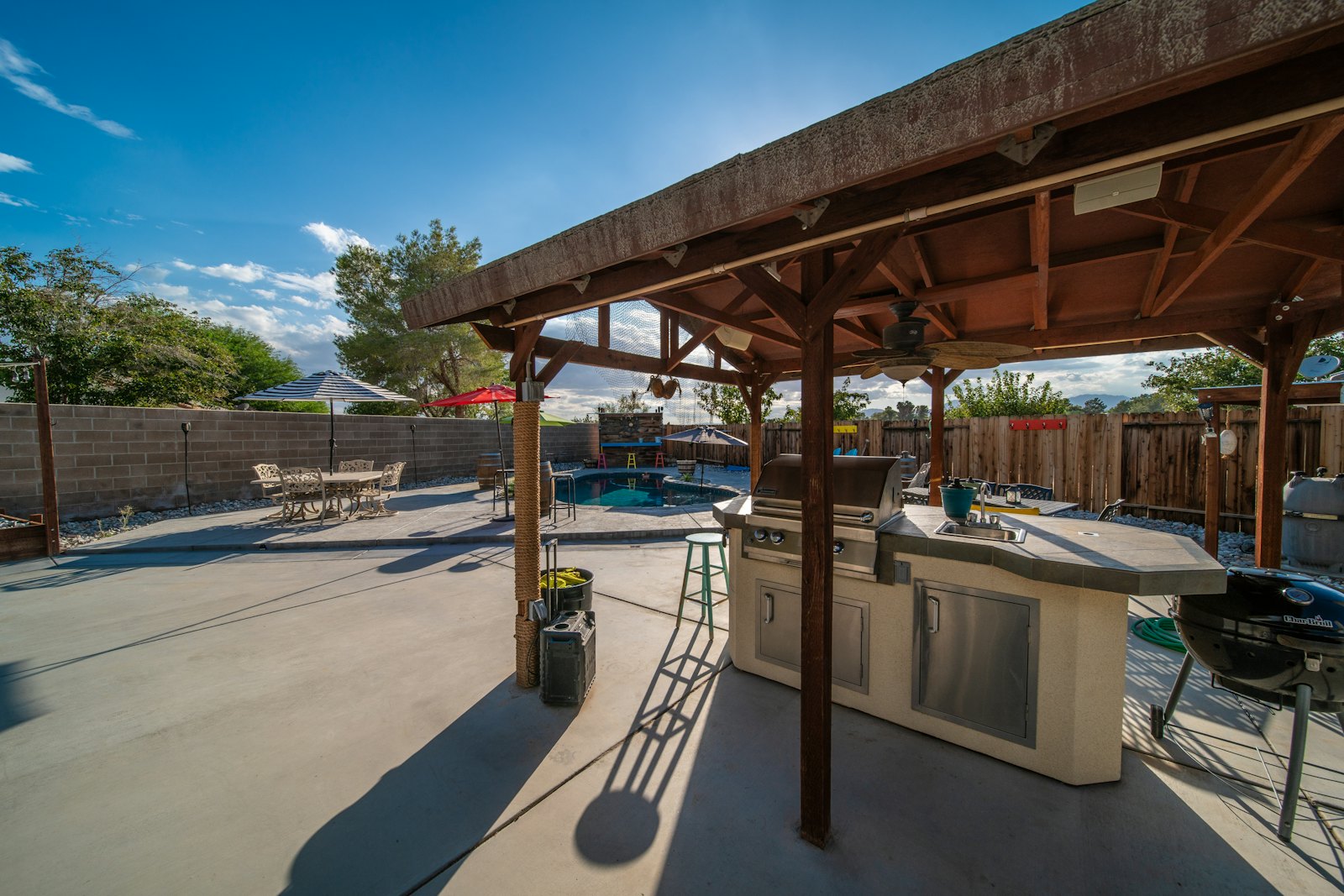Built-in outdoor kitchen and bar under a cedar pavilion on a concrete patio, with a pool in the background