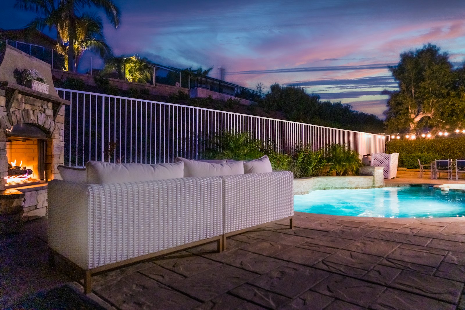 White wicker sofa beside a lit stone outdoor fireplace and pool at dusk, with string lights along the fence